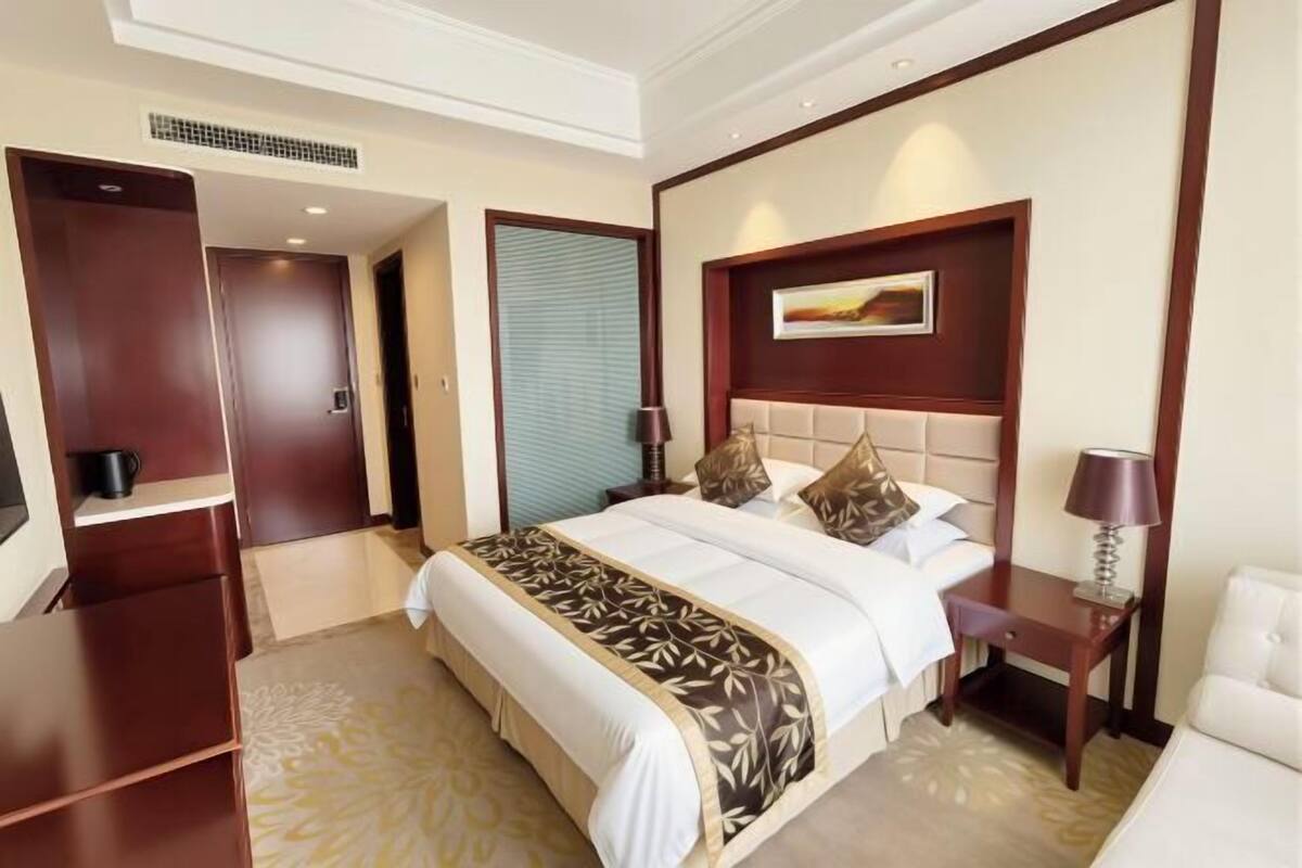 Una habitación de hotel con una cama grande, cabecera con un cuadro, una mesita de noche con lámpara y un armario.