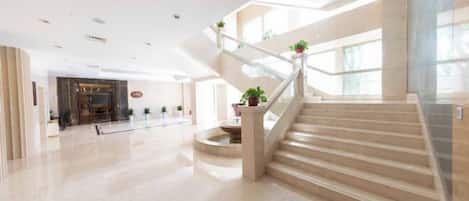 A spacious lobby with a central staircase, a reception area, and potted plants.
