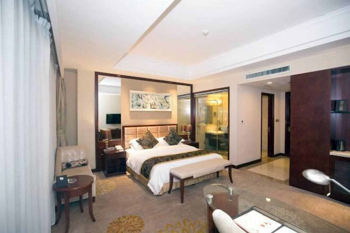 Una habitación de hotel con una cama grande, un escritorio, una silla, un sofá y un panel de vidrio.