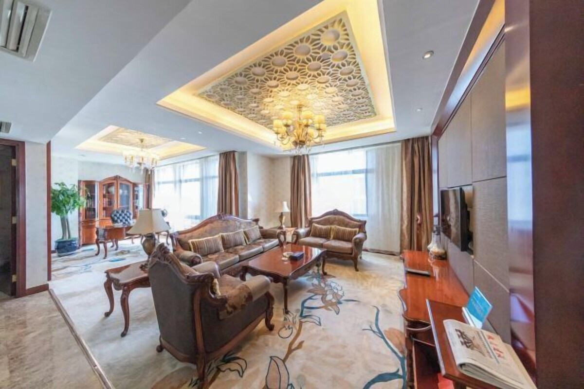 A spacious living room with a high ceiling, ornate ceiling design, and a chandelier. There is a sofa set, a coffee table, and a television on the right side.