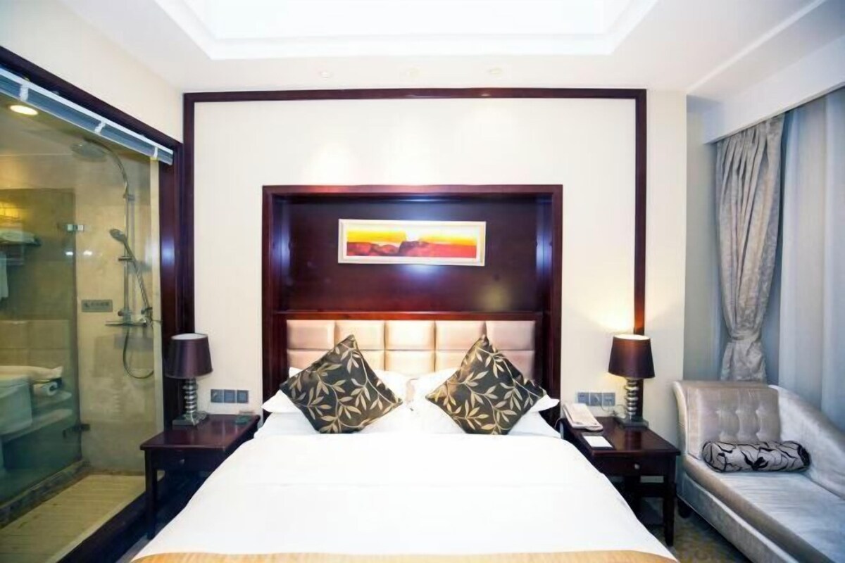 A hotel room with a large bed, a headboard with a painting, two bedside lamps, a nightstand, a sofa, and a glass-enclosed shower area.