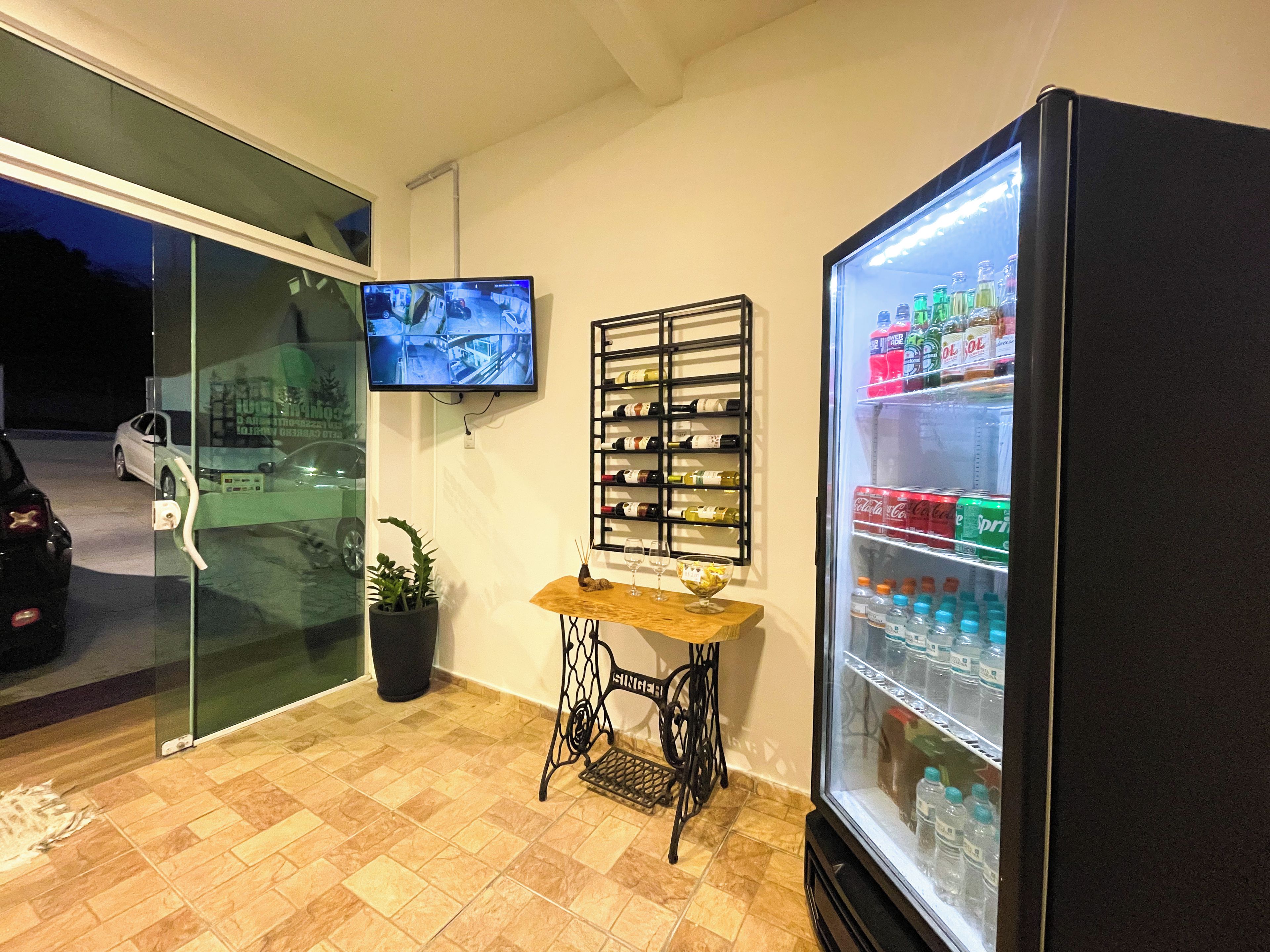 A room with a tiled floor, a television mounted on the wall, a wine rack, a refrigerator stocked with beverages, and a small table with a glass and candle.
