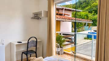 Une chambre d’hôtel avec un lit, un bureau, une chaise, une plante en pot et une vue sur une piscine et la verdure environnante.