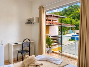 A hotel room with a bed, a desk, a chair, a potted plant, and a view of a swimming pool and greenery outside. - Pousada Mel e Mar (Penha)