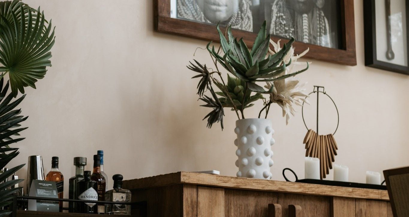 A wooden cabinet with a white vase containing plants, a framed picture on the wall, and various bottles on the left.