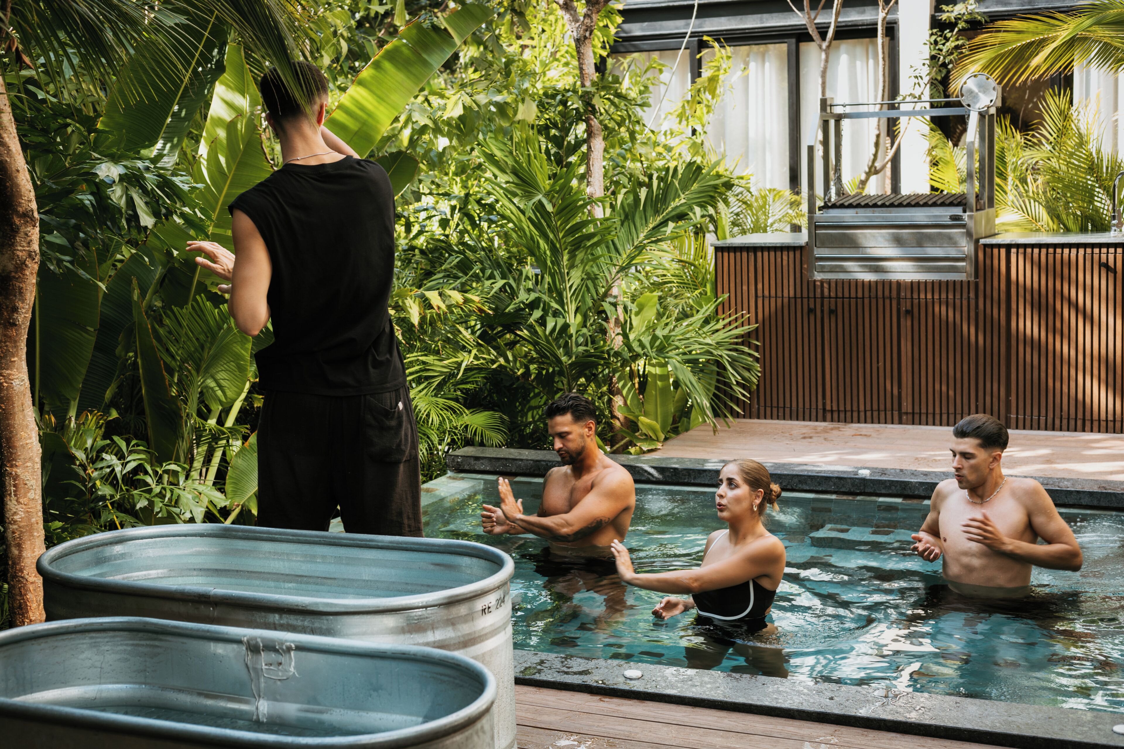 Zona de piscina con una persona junto a la piscina, tres personas en el agua y dos tinas metálicas grandes.