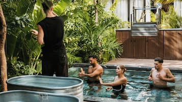 A swimming pool area with a person standing by the pool, three people in the water, and two large metal tubs.
