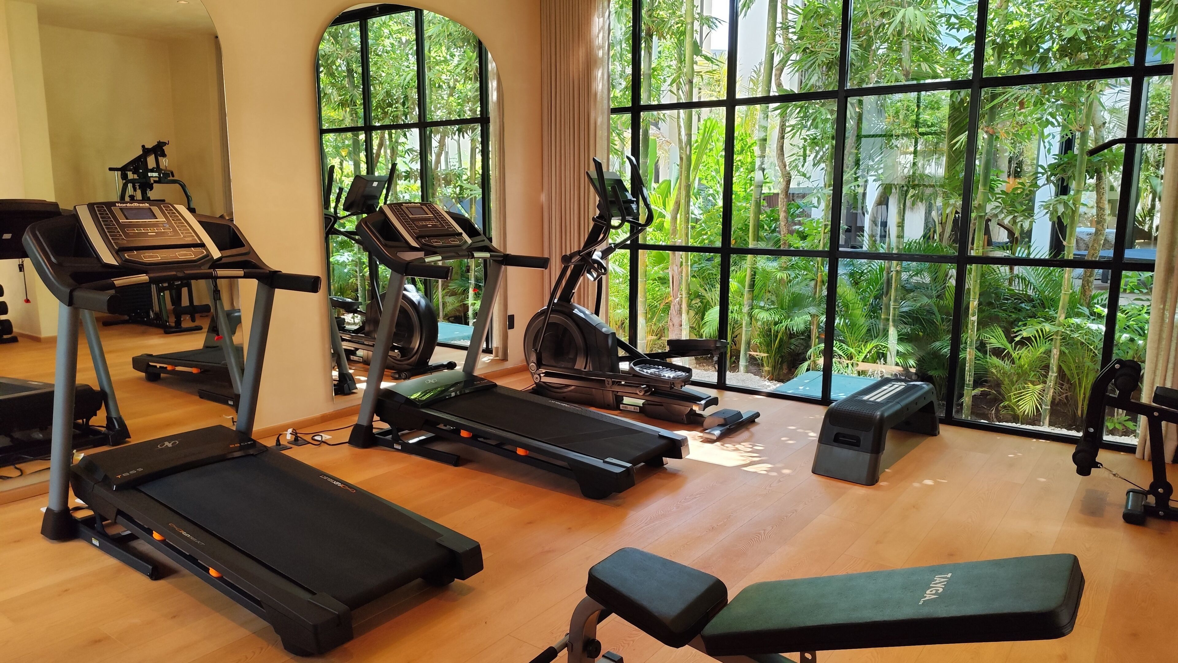 A gym with treadmills, an elliptical, and a bench, featuring large windows with a view of greenery.