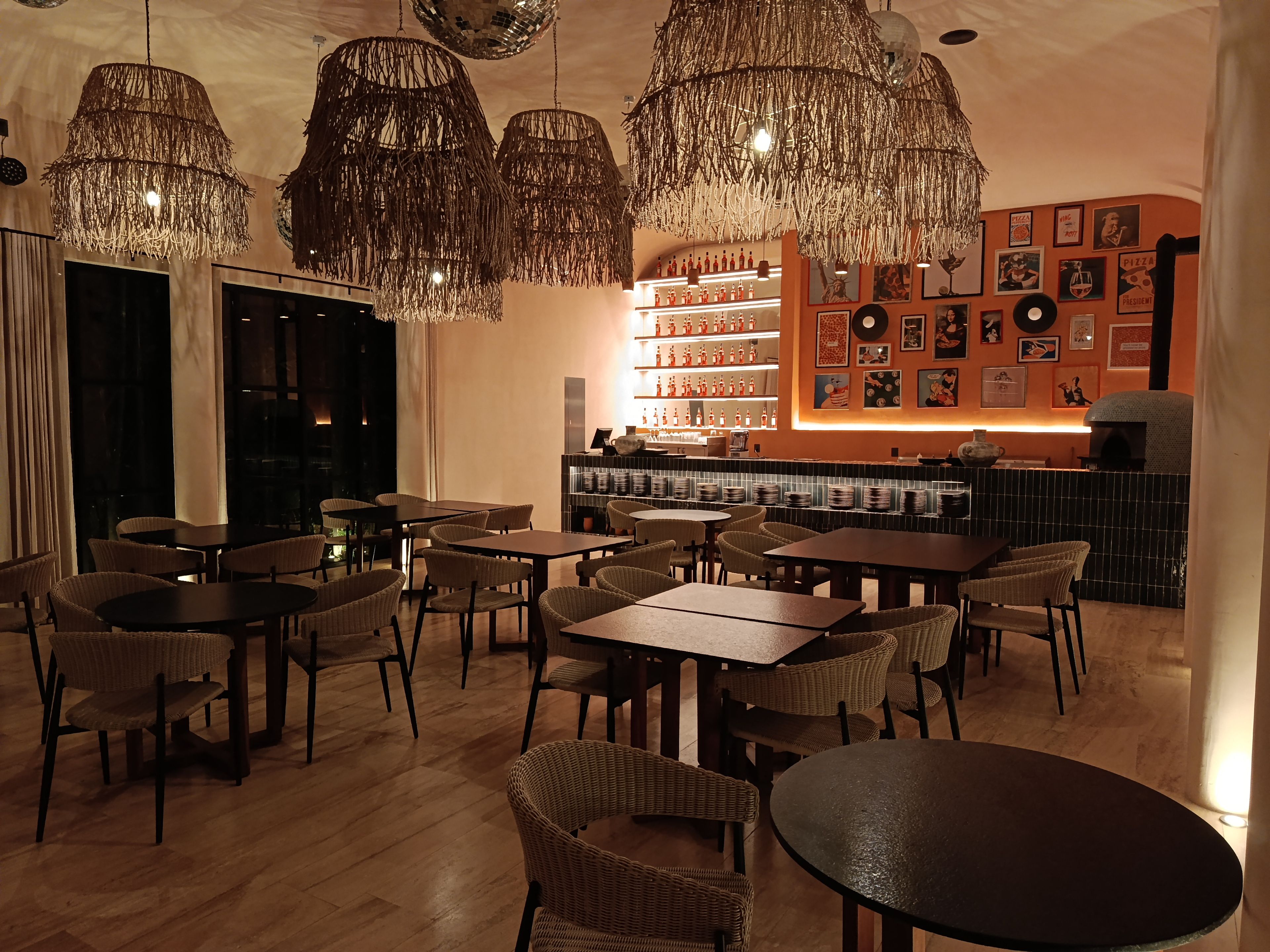 A restaurant with wicker chairs, tables, hanging chandeliers, and a bar area with shelves of bottles.