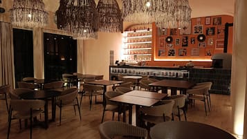 A restaurant with wicker chairs, tables, hanging chandeliers, and a bar area with shelves of bottles.
