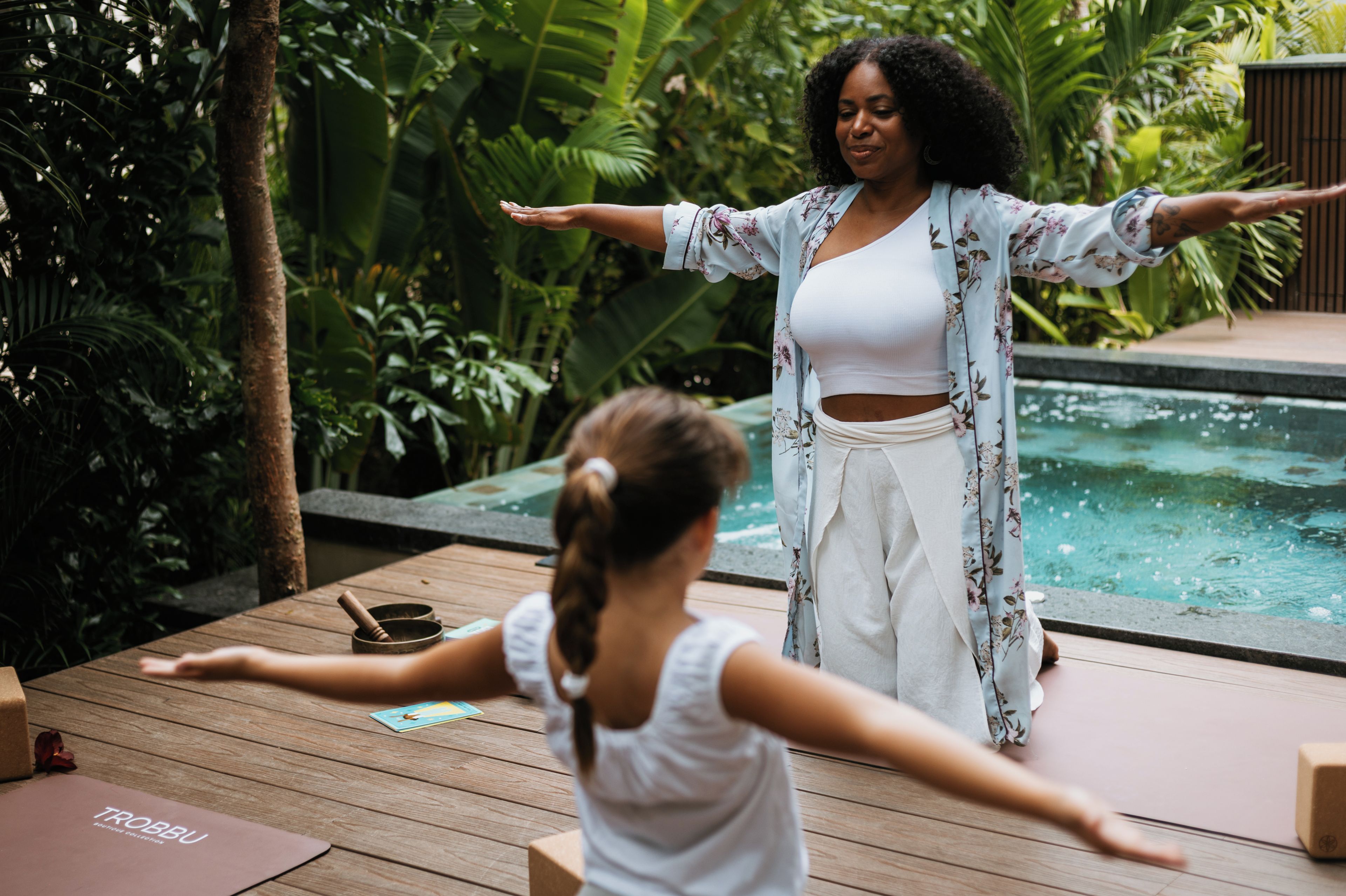 Una donna e un bambino praticano yoga all'aperto, con una piscina e vegetazione lussureggiante sullo sfondo.