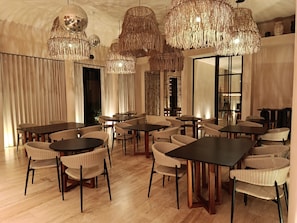 Ein geräumiger Essbereich mit mehreren Tischen und Korbstühlen, großen, hängenden Kronleuchtern und einem modernen Interieur.