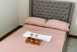 Ein ordentlich bezogenes Bett mit rosa Bettwäsche, ein grauer gepolsterter Kopfteil und ein Tablett mit zwei Gläsern und einer Flasche.