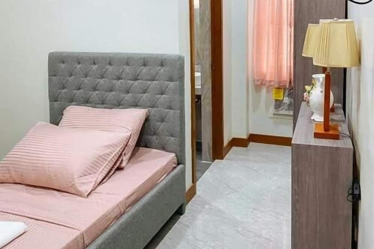 Une chambre à coucher avec une tête de lit rembourrée de couleur grise, un lit avec une literie rose, une table de chevet avec une lampe, et une vue sur la salle de bain à travers une porte en verre.