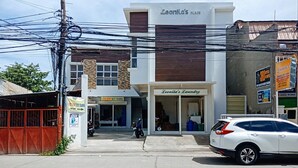 Ein Gebäude mit einem Schild, auf dem „Leonila's Place“ und „Leonila's Laundry“ steht. Vor dem Gebäude ist ein Motorrad abgestellt, und rechts daneben ein weißes Auto.