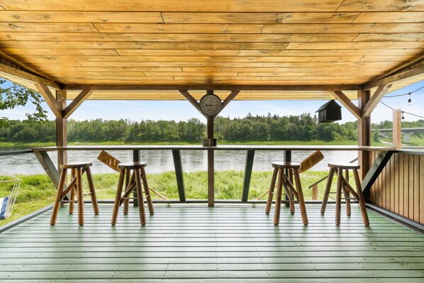 Un molo in legno con ringhiera affacciato su un fiume, con tre sgabelli di legno e un orologio.