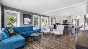 Een moderne woonkamer met een blauwe bank, een salontafel en witte stoelen. Er is een keukenhoek met witte kasten en roestvrijstalen apparatuur.