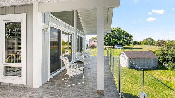 Een overdekt buitengebied met terras, stoelen en uitzicht op een grasveld en een klein gebouw.
