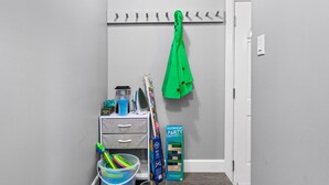Een gang met een grijze muur, een witte deur, een kapstok met een groen jasje, een kleine opbergkast met lades, een emmer met schoonmaakspullen en een kinderspeelset.