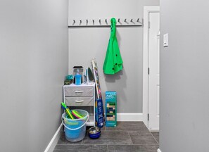 Een gang met een grijze muur, een witte deur, een kapstok met een groen jasje, een kleine opbergkast met lades, een emmer met schoonmaakspullen en een kinderspeelset.