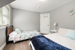 Une chambre avec deux lits, une porte blanche, une table de chevet avec une lampe et une fenêtre avec des stores.