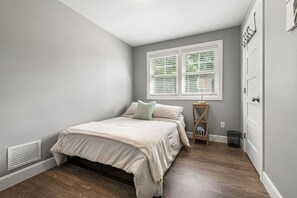Een slaapkamer met een bed, een raam met zonwering, een nachtkastje en een deur.