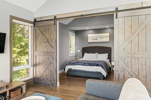 Een slaapkamer met een groot bed, een schuifdeur in boerderijstijl, een raam met zonwering en een schilderij aan de muur.