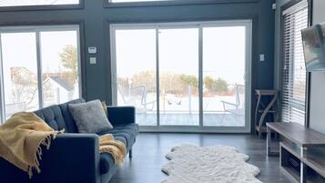 Un salon moderne avec un canapé foncé, un tapis en fausse fourrure blanche et une grande porte-fenêtre coulissante donnant sur un espace extérieur.