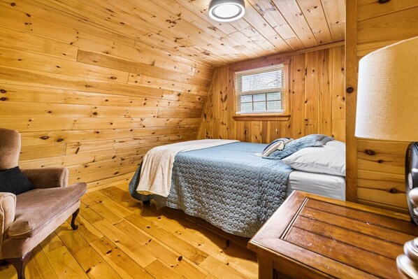 A cozy cabin room with a wooden interior, a bed with a blue quilt, a chair, and a small wooden table.