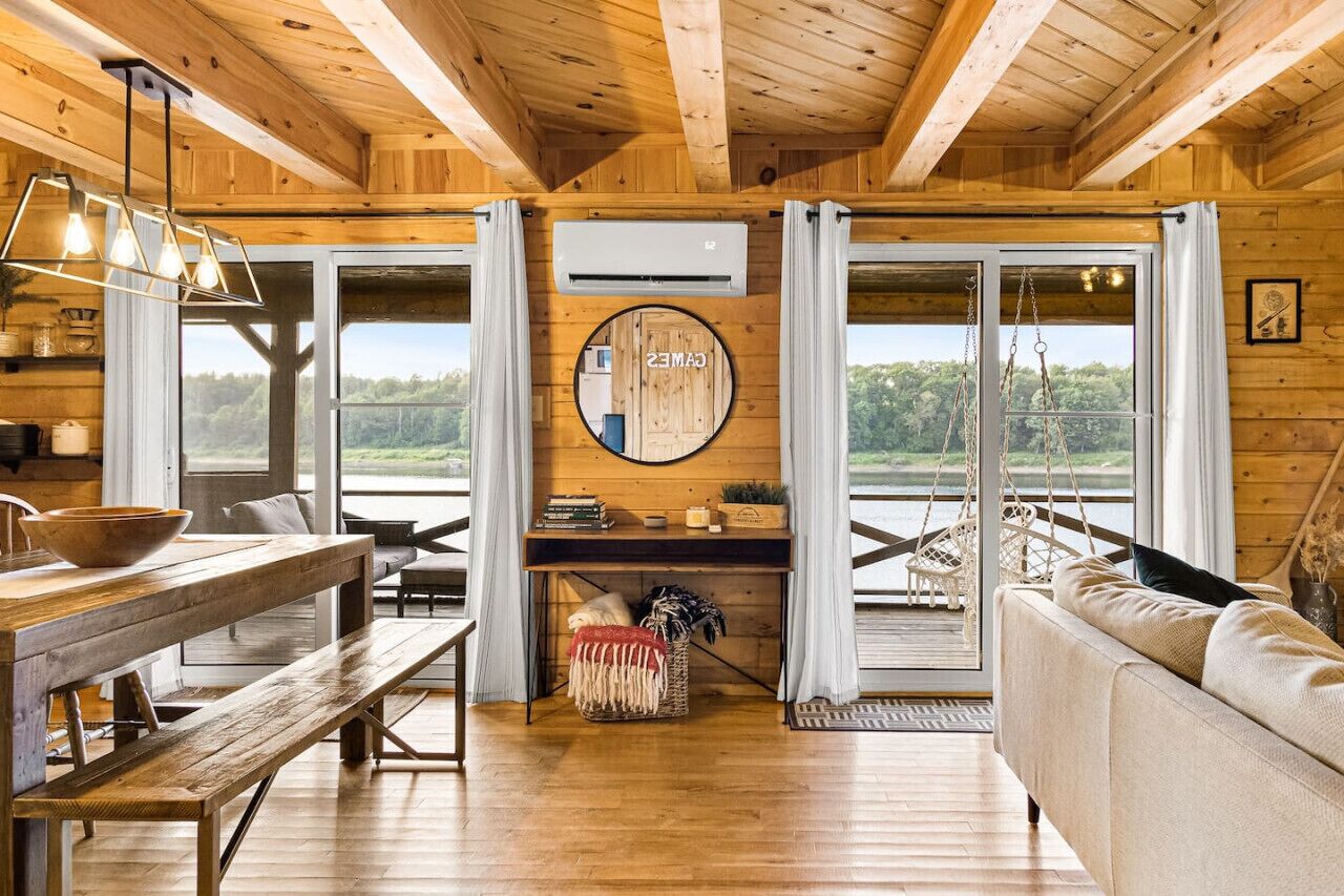 A wooden cabin interior with a dining table, bench, sofa, and large windows offering a view of the outdoors.