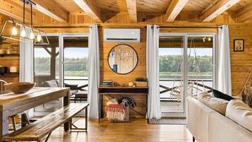 A wooden cabin interior with a dining table, bench, sofa, and large windows offering a view of the outdoors.