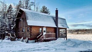 A wooden cabin with a porch and a chimney, surrounded by snow and trees.