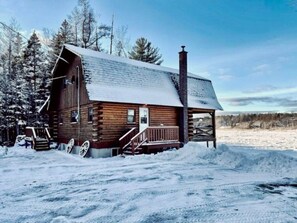 Una cabaña de madera con un porche y una chimenea, rodeada de nieve y árboles.