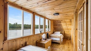 Una cabaña con paredes y suelos de madera, dos camas, una pequeña mesa con un ventilador y ventanas grandes que ofrecen vistas al exterior.