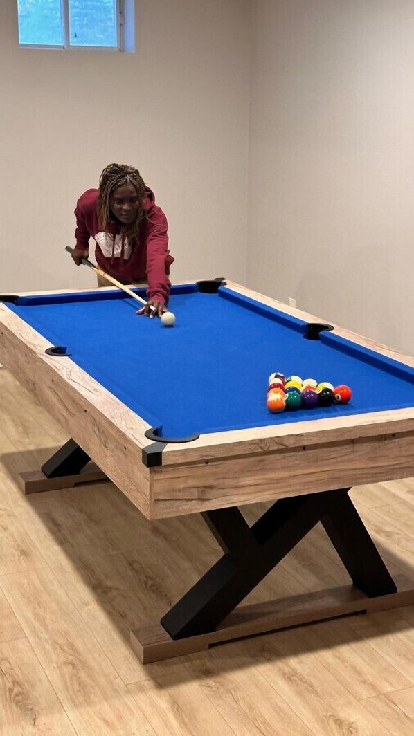 A pool table with a blue felt surface and a wooden frame, accompanied by a person in a red hoodie playing. - Richibucto River Getaway (Bass River)