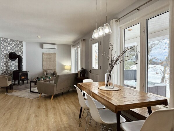 A modern living room with a fireplace, a dining area with a wooden table and chairs, and large windows offering a view of the outdoors. - Richibucto River Getaway (Bass River)