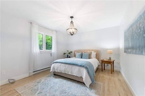 Une chambre à coucher comprenant un lit, une table de chevet, une lampe, une fenêtre avec des rideaux et un luminaire au plafond.