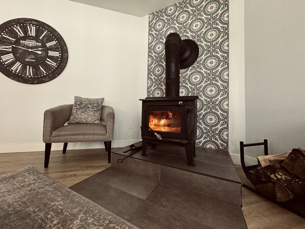 A living room with a black wood-burning stove, a grey sofa, and a patterned wall. - Richibucto River Getaway (Bass River)