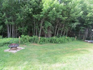 A grassy backyard with a fire pit and dense trees in the background. - Get-A-Way Beach Chalet (Grande-Digue)