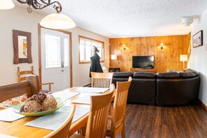 A cozy living room with a wooden wall panel, a television, a dining area with a table and chairs, and a sofa.