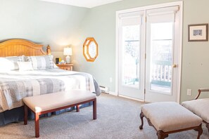 A bedroom with a large bed, a bench, a chair, a nightstand with a lamp, and a door leading to a balcony. - Get-A-Way Beach Chalet (Grande-Digue)