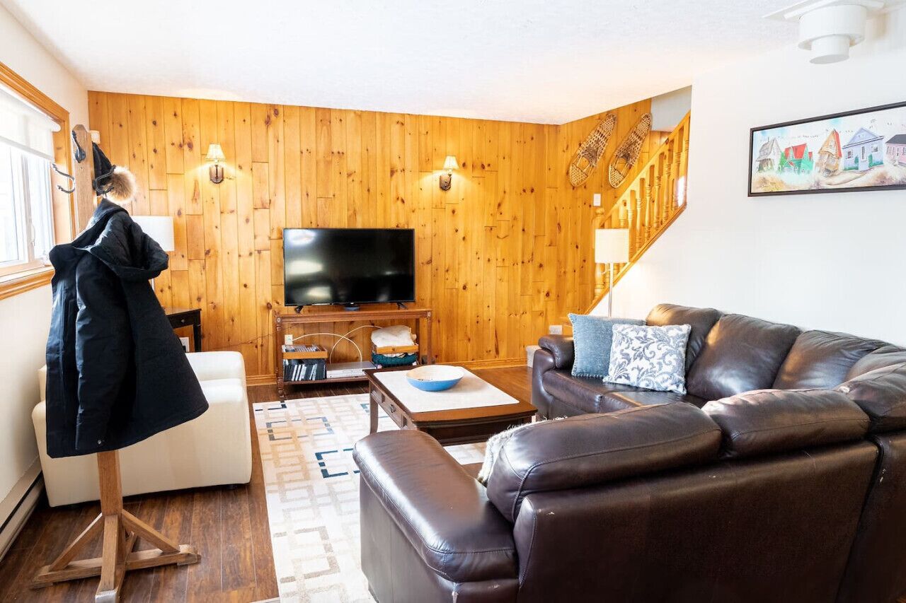 A cozy living room with a leather sofa, a flat-screen TV, wooden walls, and a staircase.