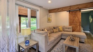 Una sala de estar con un sofá seccional color beige, una mesa de centro de madera, una lámpara y vistas al exterior con vegetación.