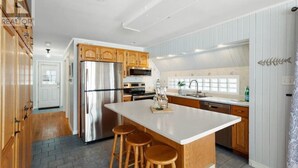 A modern kitchen with wooden cabinets, a stainless steel refrigerator, and a large island with stools. - East Coastal- Waterfront Cottage (Beaubassin-est)