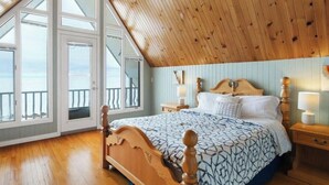 A bedroom with a sloped ceiling, a bed with a patterned bedspread, wooden headboard, bedside tables with lamps, and a view through large windows. - East Coastal- Waterfront Cottage (Beaubassin-est)