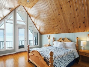 Une chambre avec un plafond en pente, de grandes fenêtres, un lit en bois et une table de chevet avec une lampe.