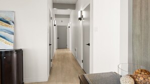 Un couloir avec un plancher en bois clair, des murs blancs et un meuble en bois foncé sur le côté gauche. Un tableau est accroché au mur et un vase en verre contenant des objets décoratifs se trouve sur le côté droit.