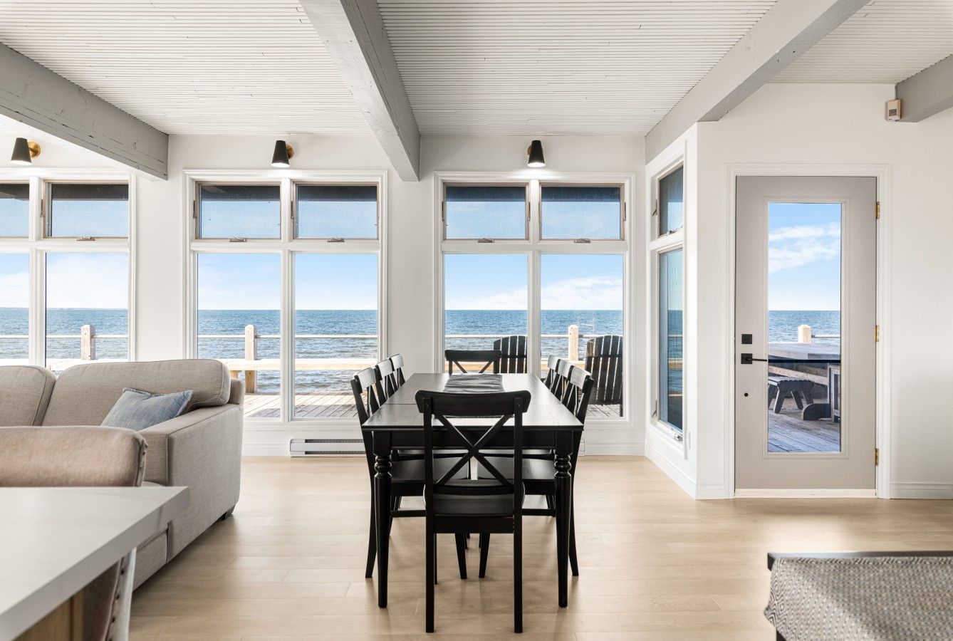 A spacious room with a large dining table and chairs, a sofa, and large windows offering a view of the sea.