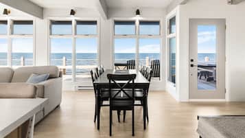 A spacious room with a large dining table and chairs, a sofa, and large windows offering a view of the sea.
