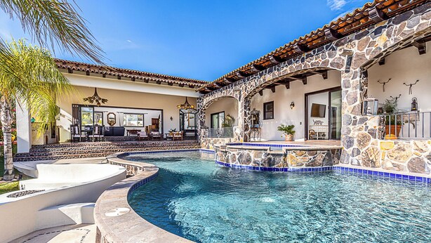 A swimming pool with a stone and tile border, surrounded by a patio area with outdoor furniture and a stone wall.